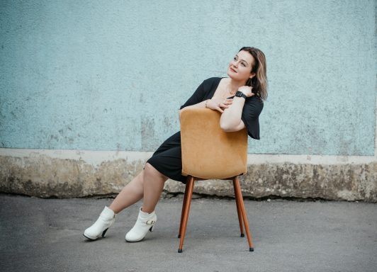 Frau in schwarzer Kleidung sitzt lässig auf einem Stuhl vor einer blauen Wand.